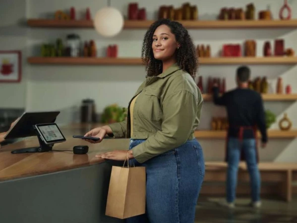 Woman paying at store with mobile phone. Customer holding shopping bag wearing jeans and jacket, purchasing goods in a shop.